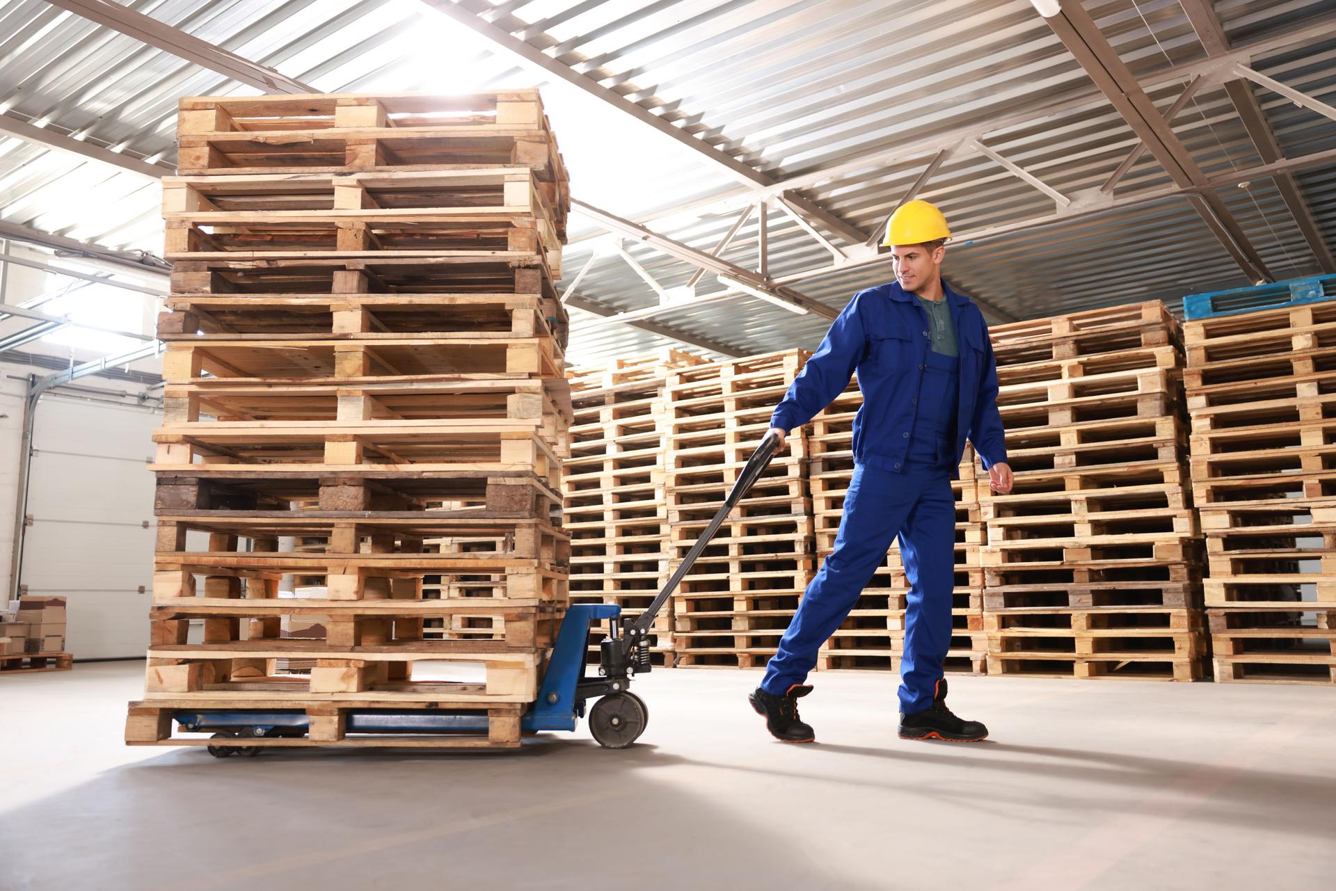 Worker moving wooden pallets with manual forklift in warehouse